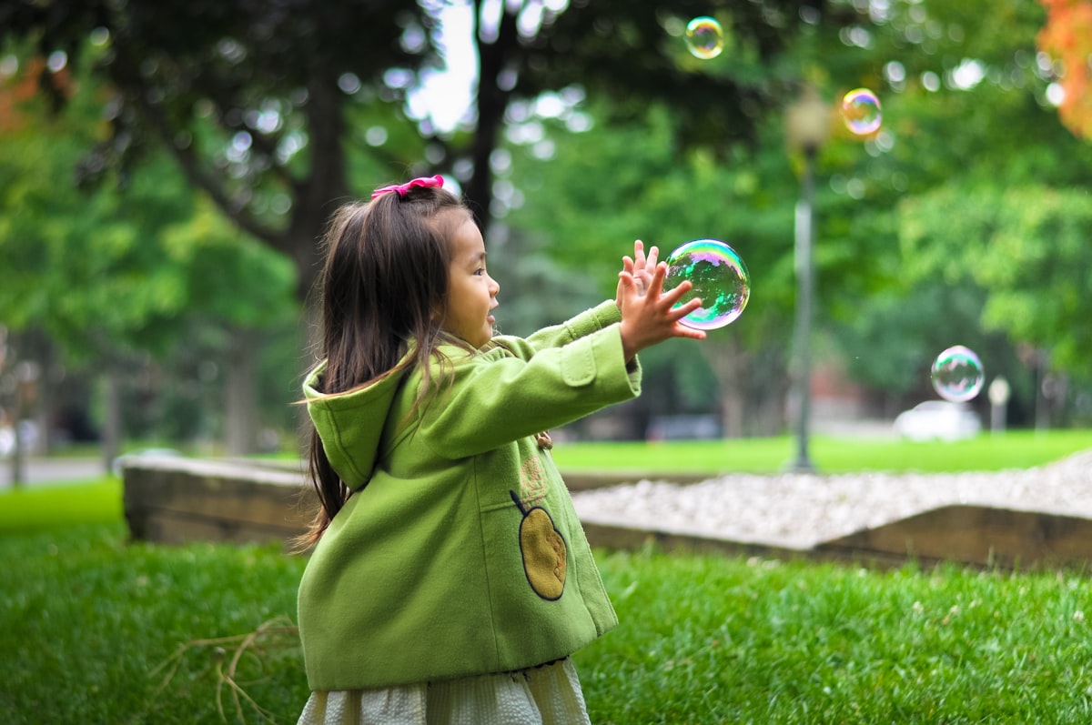 Toddler girl in a green coat catching bubbles outdoors