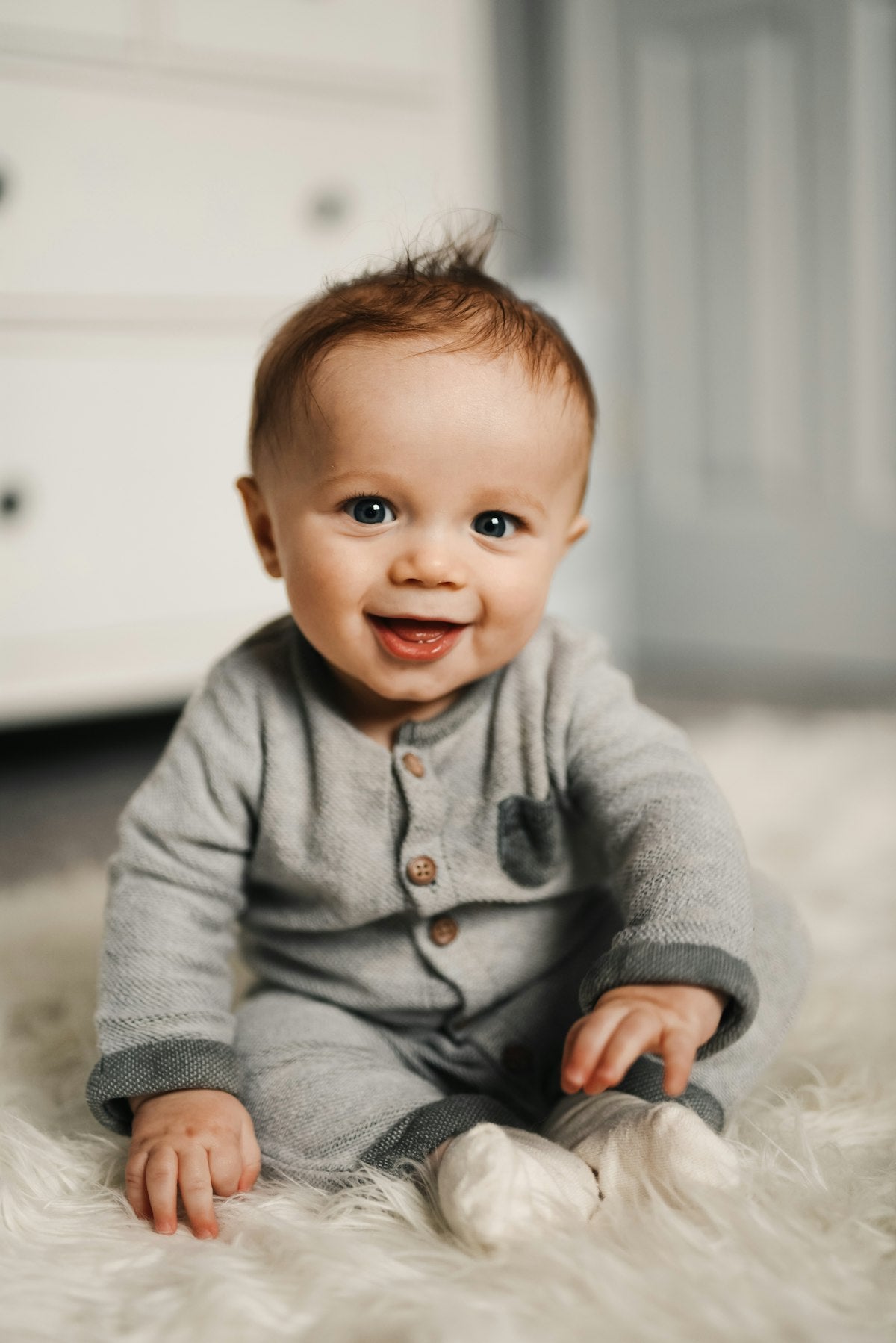 Baby in a gray knit sweater laughing and smiling, sitting on a fluffy rug